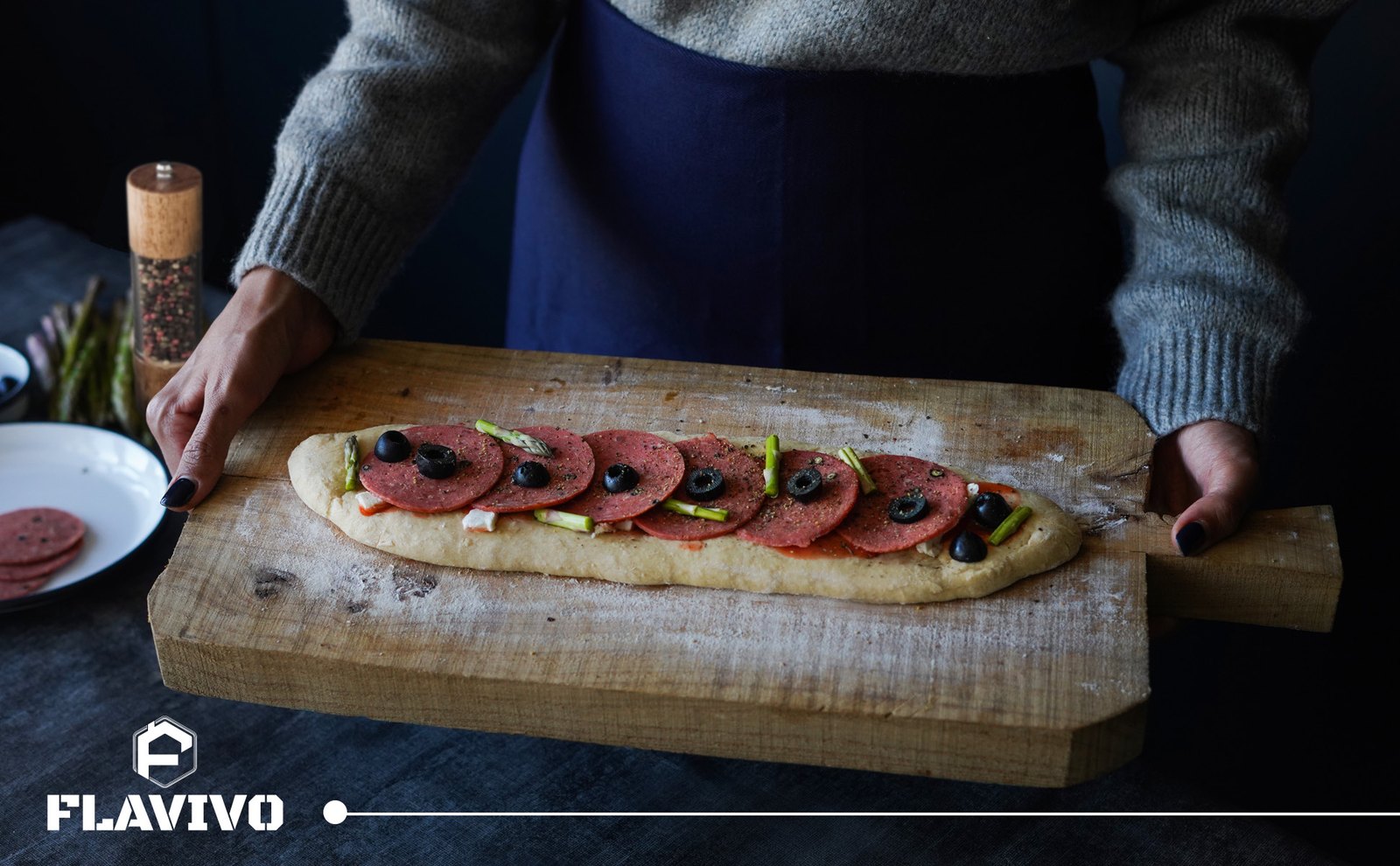 Cook holding a board with flatbread dough, a FLAVIVO grinder on the table behind